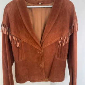 Brun mockajacka med fransar - Säljer en brun mockajacka med snygga fransdetaljer längs axlarna och ryggen. Jackan har två knappar framtill, bred krage och två fickor. Perfekt för dig som gillar western vibes och vill sticka ut med en unik look.