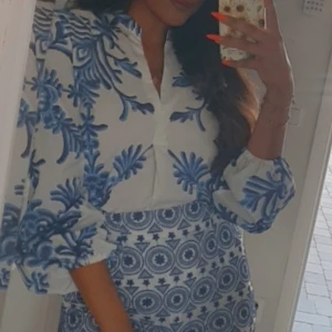 Vit och blå Positano-blus - Säljer en superfin vit och blå Positano-blus med unikt mönster. Blusen har långärmade puffärmar och v-ringad hals. Materialet känns lätt och luftigt, perfekt för varma dagar. Mönstret är både blommigt och geometriskt, vilket ger en cool och fräsch vibe.