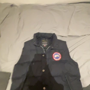 Svart dunväst från Canada Goose - Svart dunväst från Canada Goose med klassisk rund logotyp på bröstet. Västen har hög krage, tryckknappar framtill och en stilren, puffad design. Perfekt för kalla dagar när du vill hålla värmen och ändå se bra ut!
