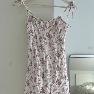 Vit blommig sommarklänning Hollister XS - Supersöt vit klänning från Hollister med rosa blommor och gröna blad. Kort modell med smala axelband som knyts upptill för en extra gullig touch. Perfekt för varma dagar och har en feminin, lätt passform. Materialet är mjukt och skönt bomull. Använd 1 gång. Säljer även liknande med blåa mönster istället 