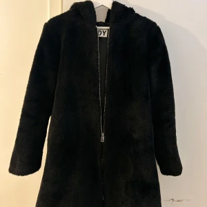 Svart teddyjacka med huva  - Mysig svart teddyjacka med huva och hel dragkedja framtill. Jackan har lång ärm och är tillverkad i mjukt pälsliknande material som ger en cool och avslappnad vibe. Perfekt för dig som gillar streetstyle och vill hålla dig varm under höst och vinter. Den har även fickor.
