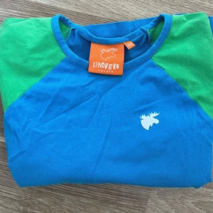 Blå och grön långärmad tröja Lindberg - Snygg långärmad tröja från Lindberg i blått med gröna ärmar. Tröjan har rund hals och ett vitt älgtryck på bröstet. Tillverkad i mjuk bomull, perfekt för chill dagar eller när du vill ha en färgglad look.