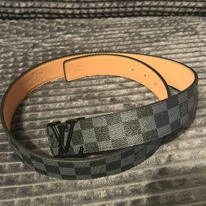 Louis Vuitton rutig skinnbälte svart - Snyggt Louis Vuitton bälte i svart och grått med klassiskt rutigt Damier-mönster. Bältet har en stilren LV-logga i svart metall som spänne och är tillverkat i exklusivt skinn. Perfekt accessoar för att lyfta din outfit med lyxig känsla.