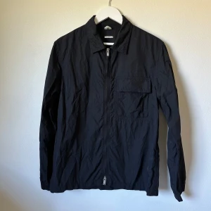 Svart vindjacka från Arket, strl 46 - Svart vindjacka från Arket i regular fit, storlek 46. Jackan har en stilren design med krage, bröstficka med lock och hel dragkedja framtill. Lätt och smidig polyester som passar perfekt när du vill ha en clean look. Långärmad och enkel att matcha med dina favoritplagg. 1A1
