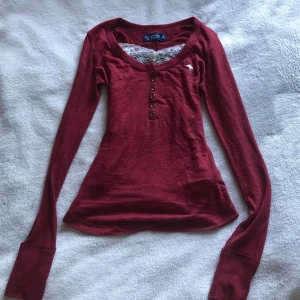 Vinröd långärmad tröja Abercrombie - Snygg vinröd långärmad tröja från Abercrombie & Fitch med rund halsringning. Tröjan har knappar framtill och en liten vit logga broderad på bröstet. Passformen är slim och materialet känns mjukt och skönt. Vit spets ingår ej.