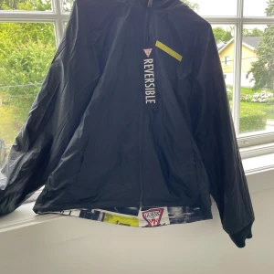 Svart och mönstrad vindjacka från Guess - Säljer en reversibel vindjacka från Guess med svart utsida och gul textdetalj, samt en insida med coolt fotomönster i svart, vitt och gult. Jackan har huva, dragkedja och ribbade muddar. Perfekt för dig som vill sticka ut med streetstyle.