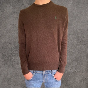 100% merinoull tröja Polo Ralph Lauren - Mörkbrun stickad tröja från Polo Ralph Lauren i merinoull storlek S. Klassisk rund halsringning och långärmad modell med broderad grön logga på bröstet. Perfekt för dig som gillar stilren och enkel design med premiumkänsla.