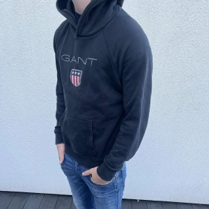 Svart hoodie från GANT med logga - Svart hoodie från GANT med broderad logga och amerikansk sköld på bröstet. Tröjan har huva, magficka och långa ärmar. Mjuk bomullskvalitet och klassisk passform, perfekt för chill dagar. Snygg och enkel att matcha med jeans eller joggers.
