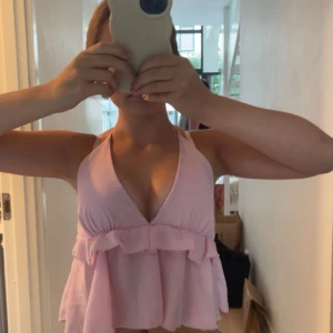 Ljusrosa volangtopp med knyt i nacken 💕 - Supersöt ljusrosa topp med volangdetalj och djup v-ringning. Toppen är ärmlös och har knyt i nacken samt öppen rygg. Perfekt för varma dagar och ger en luftig känsla. Passar dig som gillar en feminin och trendig stil.