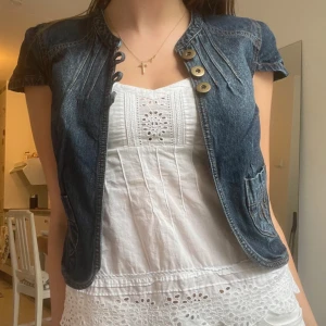 Jeansväst - Snygg jeansväst i mörkblå denim från b.p.c. Västen har korta ärmar, rund halsringning och tre stora knappar framtill. Fina sömdetaljer och två fickor framtill ger en cool och avslappnad look.