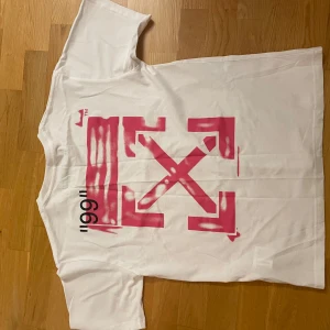 Vit Off-White t-shirt med rosa print - Säljer en vit t-shirt från Off-White med ikoniskt rosa pilar-print på ryggen och svart text framtill. T-shirten har korta ärmar, rund hals och är gjord i mjuk bomull. Snygg streetwear-vibe och coola detaljer som 'IMPRESSIONISM' tryckt på framsidan.