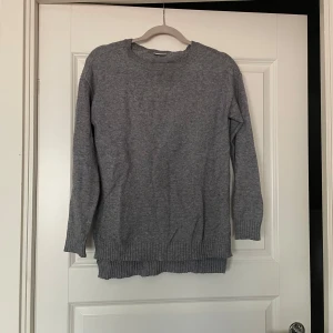 Grå stickad tröja med rund hals - Mysig grå stickad tröja med rund halsringning och långa ärmar. Tröjan har ribbade muddar vid ärmslut och nederkant, vilket ger en snygg och enkel look. Perfekt att slänga på sig när det är kyligt ute.