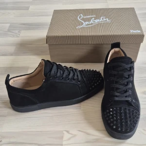 Svarta sneakers från Christian Louboutin - Snygga svarta sneakers från Christian Louboutin med klassiska röda sulor och coola nitar på tån. Skorna har snörning och är tillverkade i mocka med detaljer i skinn. Perfekta för dig som vill sticka ut med en edgy och lyxig stil.