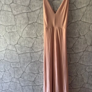 Ljusrosa långklänning med v-ringning - Säljer en elegant ljusrosa långklänning med djup v-ringning och tunna axelband. Klänningen har en flowy passform och öppen rygg med smala banddetaljer. Perfekt för dig som vill ha en stilren och feminin look. Katt finns i hemmet.
