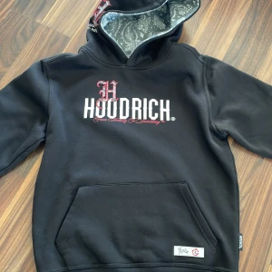 Svart hoodie från Hoodrich med paisley - Svart hoodie från Hoodrich med stor vit logga och röd detalj på bröstet. Insidan av huvan har ett snyggt paisleymönster i grått och vitt. Tröjan har magficka och tryck även på ryggen. Materialet är 80% bomull och 20% polyester, perfekt för chill dagar.