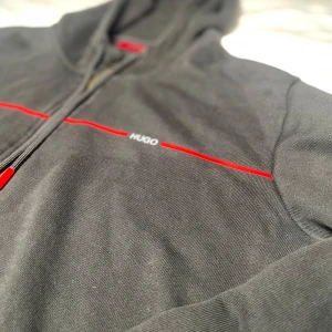 Svart hoodie från Hugo med röd detalj - Säljer en svart Hugo boss tröja då tröjan är ganska använd men ändå i bra skick den har ej några hål eller fläckar kom gärna privat för mer info eller bilder på tröjan😊