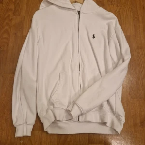 Vit hoodie från Polo Ralph Lauren - Vit hoodie från Polo Ralph Lauren i storlek L. Klassisk design med dragkedja, huva och kängurufickor. Broderad svart logga på bröstet. Mjuk bomullskvalitet och ribbade muddar för en skön passform. Perfekt för en clean och stilren look.