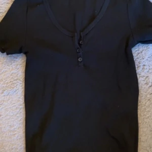 Svart kortärmad topp med knappar - En svart, enkel topp med korta ärmar och v-ringning framtill. Toppen har tre små knappar vid halsringningen som ger en snygg detalj. Perfekt att matcha med jeans eller kjol för en clean look.