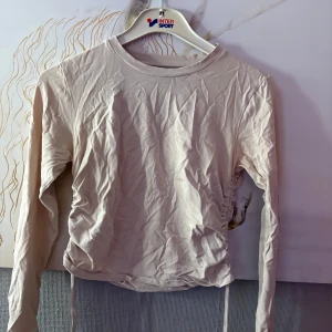 Beige långärmad topp från Pieces - Snygg beige långärmad topp från Pieces med trendig snörning i sidorna som ger en cool rynkad effekt. Tillverkad i mjuk bomull, perfekt för lager-på-lager och passar till både jeans och kjol. Enkel rund halsringning och relaxed fit.