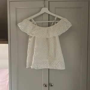 Vit blus med volang och broderi - Supersöt vit blus med axelband och bred volangkant som går offshoulder. Tunt bomullstyg med broderade hål och lätt struktur, perfekt för varma dagar. Snygg till jeans eller kjol för en fräsch sommarlook.