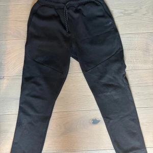 Svarta mjukisbyxor från Nike - Svarta mjukisbyxor från Nike med elastisk midja och snörning. Byxorna har dragkedjeförsedda fickor och en diskret Nike-logga på benet. Perfekta för chill eller träning, med en skön och avslappnad passform.