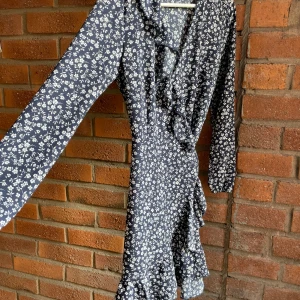 Blommig omlottklänning från ONLY - Supersöt kort omlottklänning från ONLY i marinblått med vitt blommigt mönster. Klänningen har långa ärmar, volangdetaljer längs kanten och knyt i midjan för en snygg siluett. Perfekt för dig som gillar romantisk och trendig stil.