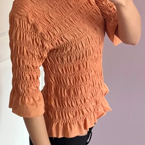 Persikofärgad blus från Gina Tricot - Supersöt blus i persikofärg från Gina Tricot med smockad struktur och volangdetaljer vid ärmslut och nederkant. Blusen har rund hals och trekvartsärm, perfekt för dig som gillar en feminin och trendig look. 