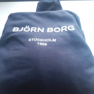 Mörkblå Björn Borg hoodie - Snygg mörkblå hoodie från Björn Borg med vit text på bröstet där det står 'BJÖRN BORG STOCKHOLM 1956'. Tröjan har en klassisk huva och är perfekt för chill dagar. Mjuk bomullskvalitet och stilren design. Utan defekter. Nypris ca 600kr