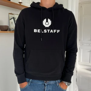 Svart Belstaff hoodie med logga - Svart hoodie från Belstaff med vit logga och text på bröstet. Tröjan har huva med snörning, stor magficka och långa ärmar. Mjuk och skön bomullskvalitet. Snygg och enkel design som passar bra till jeans.