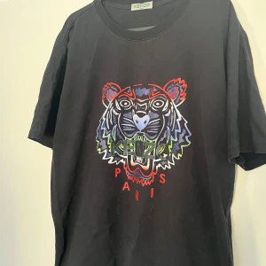 Svart Kenzo t-shirt med tigertryck - Svart t-shirt från Kenzo med ikoniskt tigertryck i flera färger på bröstet och texten 'Kenzo Paris'. Rund hals och korta ärmar. Materialet är mjuk bomull som känns skönt mot huden. Perfekt statement-plagg för dig som gillar streetstyle och coola prints.