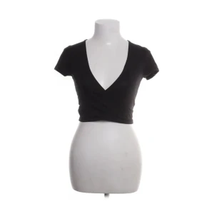 Svart croppad topp från NLY Trend - Svart croppad topp från NLY Trend med djup v-ringning och korta ärmar. Snygg och enkel design som sitter tight och framhäver midjan. Perfekt att matcha med höga jeans eller kjol för en trendig look.