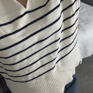 Randig stickad tröja från Zara - Säljer en snygg stickad tröja från Zara i storlek XS. Tröjan är krämvit med marinblå ränder, har en bred krage med v-ringning och långa ärmar. Ribbad nederkant och muddar ger en chill vibe. Perfekt för dig som gillar klassisk stil med en twist. Den är oanvänd.