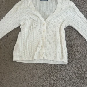 Vit kofta från Brandy Melville  - Säljer en vit kabelstickad kofta från Brandy Melville med v-ringning och knappar framtill. Koftan har långärmade ärmar och ribbade muddar vid ärmslut och nederkant. Perfekt att slänga över en topp för en chill look.