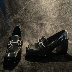 Svarta platåloafers med klack från H&M - Snygga svarta loafers i lack från H&M med chunky platåsula och hög klack. Skorna har fyrkantig tå, dekorativ metallspänne framtill och en glansig finish som ger en cool retrovibe. Perfekta för dig som vill sticka ut med din stil.
