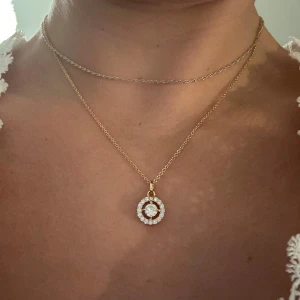 Guldigt halsband med glittrig cirkelberlock - Snyggt halsband i guld med en rund berlock dekorerad med vita stenar. Kedjan är tunn. Berlocken har en cirkelformad design med stenar både i mitten och runt om. Nypris 349kr                   Kom med prisförslag! Tryck INTE på köp nu