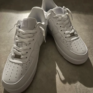 Nike Air Force 1 helvita sneakers - Klassiska Nike Air Force 1 i helvitt skinn med perforerad tå, vit sula och snörning. Snygg och tidlös modell med Nike-logga på sidan och bak. Perfekta för dig som vill ha en clean och fräsch look till vardags. Hör av vid minsta fundering 💫