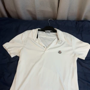 Vit Moncler pikétopp med krage - Vit pikétopp från Moncler med klassisk krage och korta ärmar. Liten broderad Moncler-logga på bröstet. Perfekt för en clean och sportig look. Materialet känns mjukt och svalt mot huden.