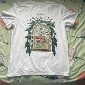 Vit Avenida Casablanca t-shirt - Vit t-shirt med coolt tryck på både bröstet och ryggen. Stort grafiskt motiv med öga, berg och valv omgivna av gröna blad. Klassisk passform och rund hals. Perfekt för dig som gillar unika prints och streetstyle.