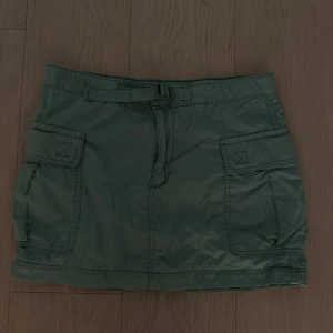 Grön cargokjol från Levi's, stl 26 - Snygg grön cargokjol från Levi's med praktiska fickor på sidorna och ett bälte i midjan. Kjolen är kort och har en avslappnad, cool stil som passar perfekt till sneakers eller boots. Materialet är lätt. Aldrig använd eller tvättad. Storlek 26. Köparen står för frakt eller mötas på Ekerö 