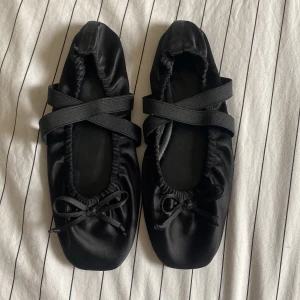 Svarta ballerinaskor med rosett - Snygga svarta ballerinaskor från ASOS i satin material med elastiska band som korsas över foten och en söt svart rosett framtil som bara använts en gång och är i mkt bra skick!🖤🤗