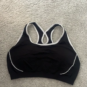 Svart sport-bh från Active Intent - Sport-bh i svart med vita kanter från Active Intent. Snygg racerback-modell med kontrastfärgade sömmar och diskret struktur på nedre delen. Tillverkad i en mix av nylon, polyester och elastan som ger bra stöd och stretch. Perfekt för träning och aktiva dagar. Har aldrig använts. 