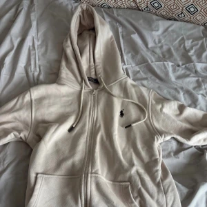 Beige hoodie från Polo Ralph Lauren - Säljer en beige hoodie från Polo Ralph Lauren med dragkedja och snörning i huvan. Klassisk broderad logga på bröstet och stora fickor framtill. Mjuk bomullskänsla och perfekt för chill dagar. Passar dig som gillar stilren och enkel look.(Pris kan diskuteras) Passar också om man har storlek M