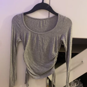 Grå ribbad topp med snörning från SHEIN - Grå långärmad topp från SHEIN i ribbat material. Modellen har rund halsringning och snygg snörning i sidorna som ger en trendig, rynkad effekt. Perfekt för dig som gillar en enkel men ändå stylish look. Sitter tajt och fint.