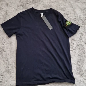 Mörkblå Stone Island t-shirt - Snygg mörkblå t-shirt från Stone Island med klassisk rund hals och korta ärmar. På vänster ärm sitter den ikoniska Stone Island-patchen med broderad logga i gult och grönt. Tillverkad i mjuk bomull för skön känsla. Perfekt för dig som gillar streetwear och stilrena plagg.