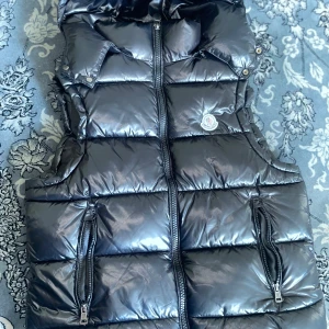 Svart dunväst från Moncler med huva - Snygg svart dunväst från Moncler med glansig finish och vadderad design. Västen har huva, två dragkedjefickor framtill och Moncler-logga på bröstet. Perfekt för lager-på-lager och ger en clean streetstyle-vibe.