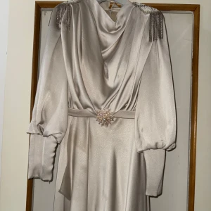 Champagnefärgad långklänning med pärldetaljer - Säljer en fantastisk champagnefärgad långklänning med en lyxig glans och elegant drapering framtill. Klänningen har vackra puffärmar, pärldekorerade axlar och glittrande fransar som verkligen fångar blickar. Den är dessutom utrustad med ett bälte prytt med en stor pärlbeströdd brosch som ger en exklusiv känsla. Köpt för 3500 kr och använd endast 5 timmar – knappt använd och i toppskick! Perfekt för dig som vill glänsa och göra ett starkt intryck på festen.