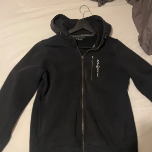 Svart hoodie från Sail Racing - Svart hoodie från Sail Racing med dragkedja framtill och bröstficka med logga. Hoddien har huva och är långärmad, perfekt för dig som gillar en clean och sportig stil. Materialet känns mjukt och bekvämt, och passformen är normal.