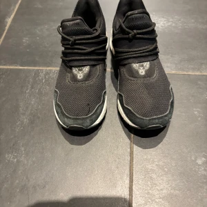 Svarta Plein Sport sneakers med silverdetalj - Svarta sneakers från Plein Sport med mesh- och mockadetaljer, snörning och en cool silverfärgad häl. Skorna har vit sula och ett stilrent tryck av ett tigerhuvud på tungan. Perfekta för dig som gillar sportig och modern stil.