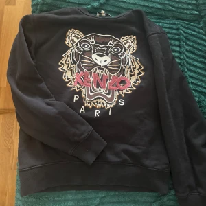 Svart Kenzo sweatshirt  - Svart sweatshirt från Kenzo i bomull med ikoniskt broderat tigerhuvud och logga i rosa och beige på bröstet. Tröjan har rund hals, långa ärmar och en relaxed passform. Perfekt statement piece för dig som gillar streetstyle.