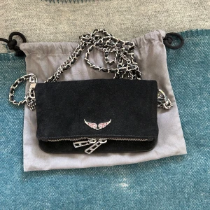 Svart mocka axelväska Zadig & Voltaire - Supersnygg svart axelväska i mocka från Zadig & Voltaire! Endast använd en gång och är köpt av mig från NK! Framsidan har en liten vingdekor med rosa stenar. Perfekt storlek för det viktigaste, gör verkligen en outfit! Pris kan diskuteras, då den inte kommer till användning av mig🤗